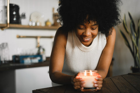 A smiling woman with hands cupped around a medium candle. Sourced from Pexels.
