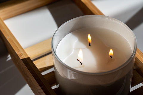 A large three-wick candle that is lit, positioned on a wooden bath rack