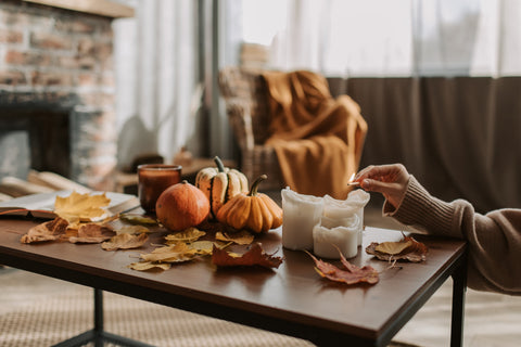 A woman lighting a candle positioned on an autumnal display featuring pumpkins and leaves. Sourced from Pexels.