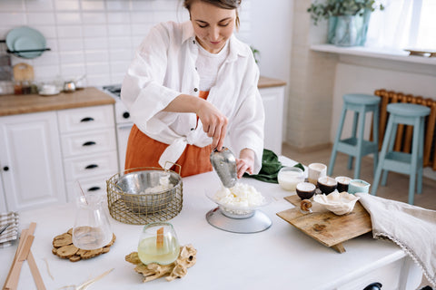 A smiling woman making candles at home, using a scoop to organise the candle wax. Sourced from Pexels.