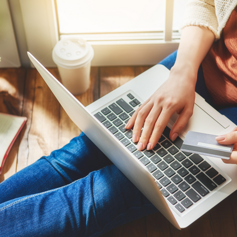 someone is sitting on the floor with a laptop in their lap. Typing with one hand and a credit card in the other