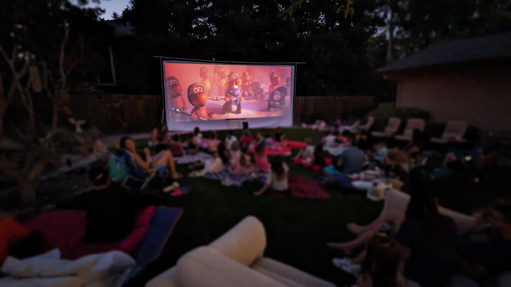 A bunch of children and adults sitting on the ground and watching an outdoor movie