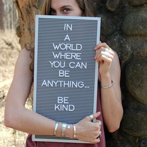 Photo of a young woman holding a message board that reads "In a World Where yOU Can be anything, be kind" Young woman is wearing Upcycled silverware jewelry