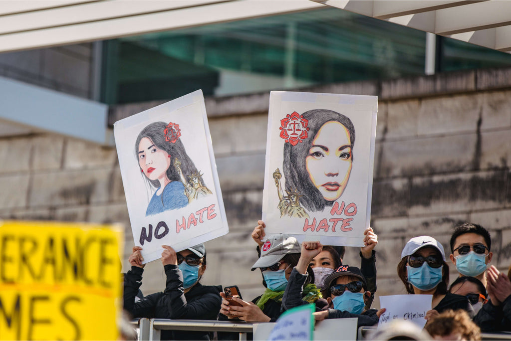 Two activists at a #StopAAPIHate rally holding up white signs with illustrations of AAPI women, the Statue of Liberty, a red flower symbol with a balanced scale at the center, and "No Hate" written on the bottom.