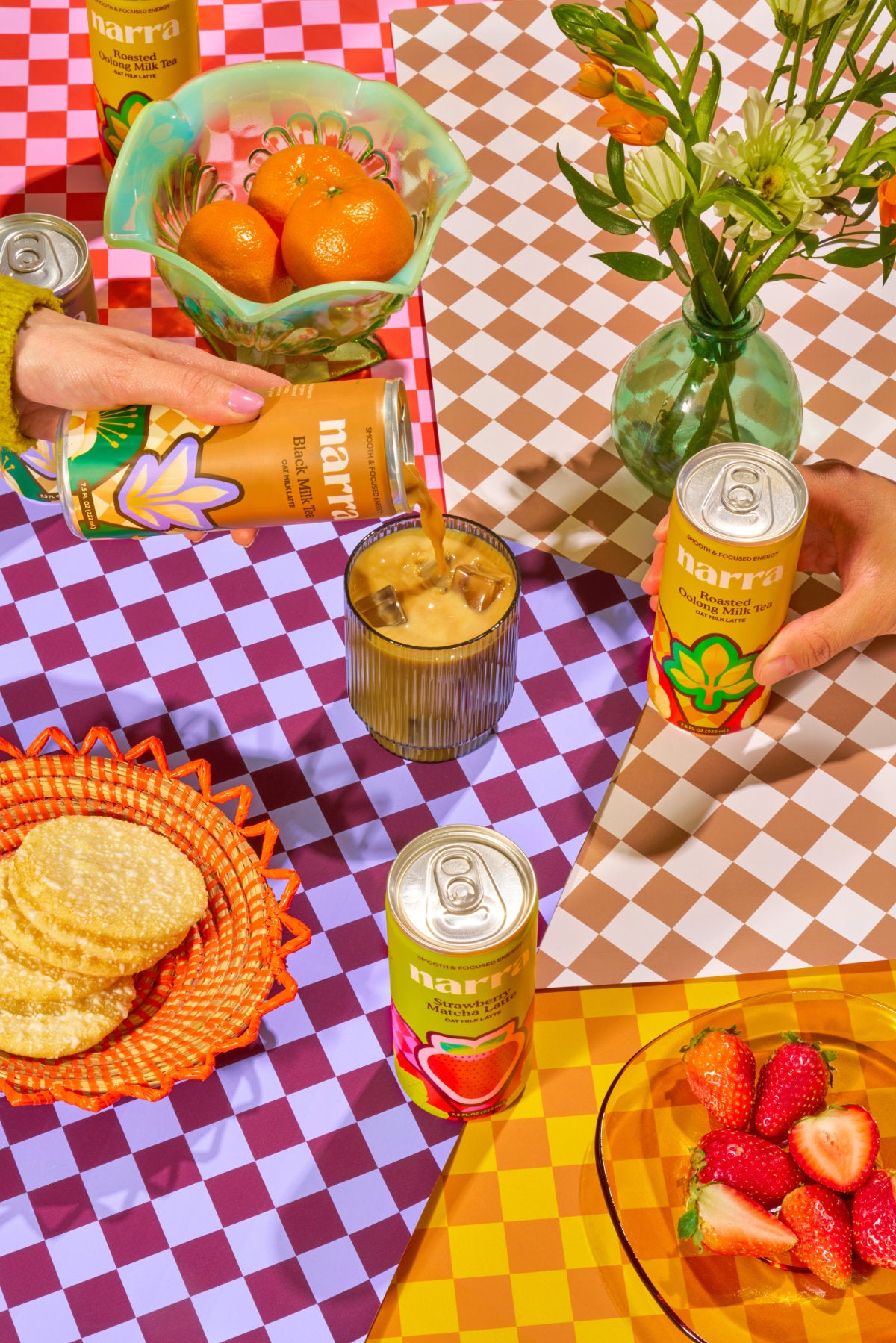 Narra's oat milk lattes on a table with strawberries, cookies, oranges, and flowers, being poured into glasses.