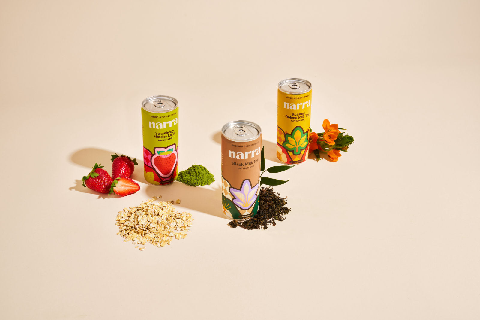 Narra's oat milk lattes on a beige background with ingredients sprinkled around the cans: matcha, loose tea, oats, strawberries, and flowers.