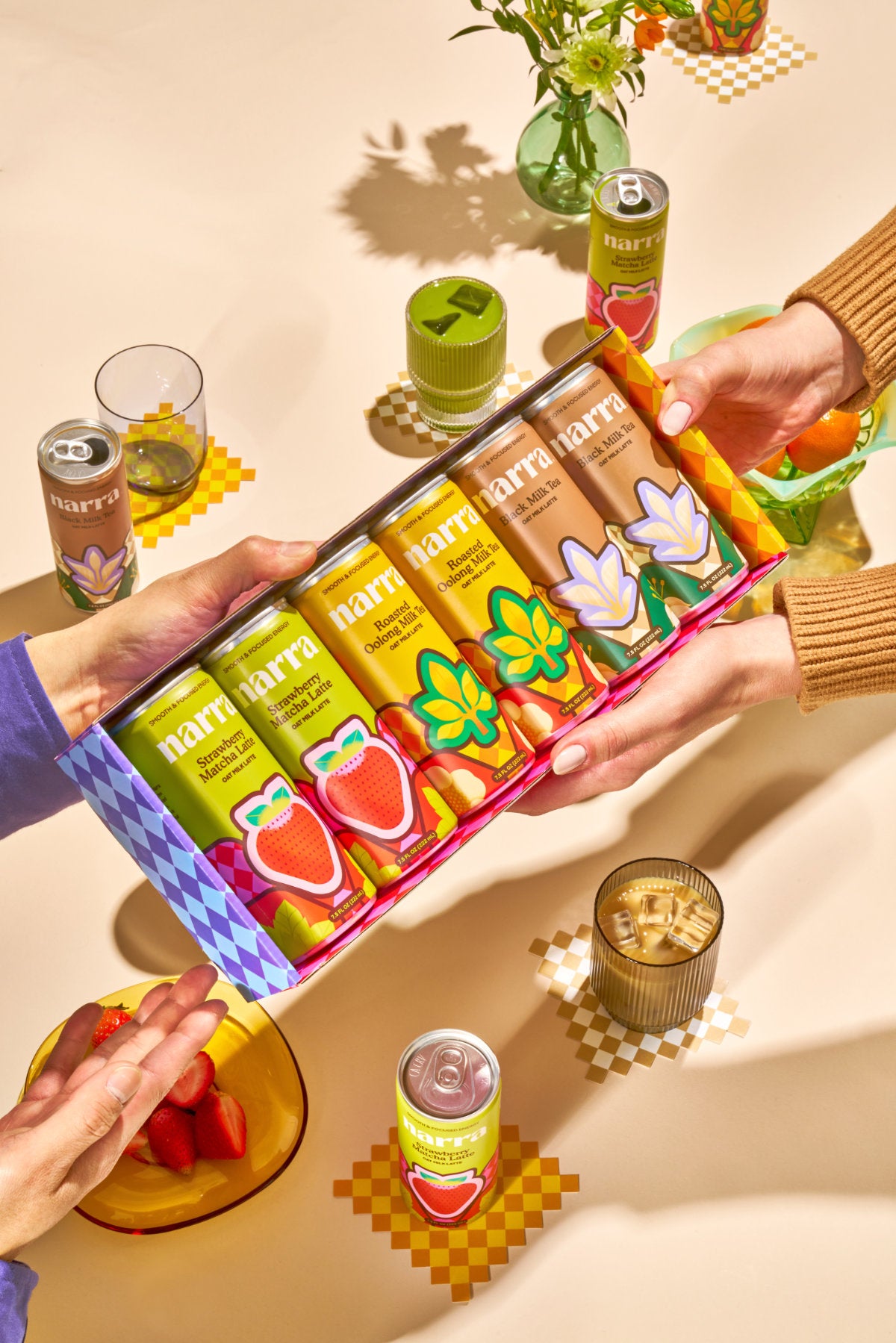 Two pairs of hands holding up an open Narra oat milk latte variety pack, showcasing six cans, above a table with the oat milk lattes in cans and poured into glasses.