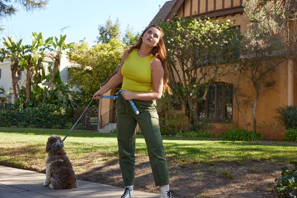 A woman makes a face as she tugs on a leash and her curly dog sits stubbornly on the sidewalk.