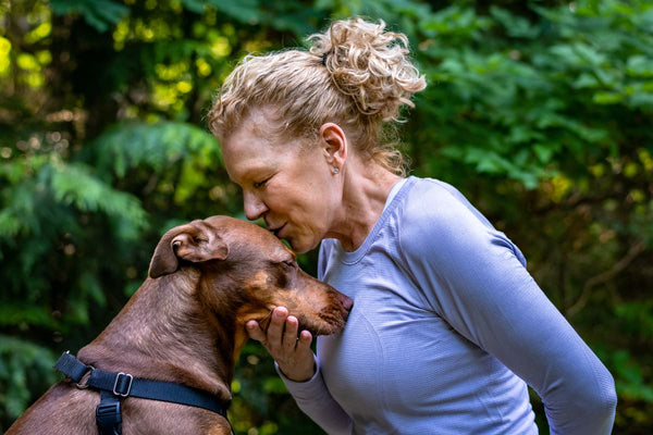 A woman with blond curly hair kisses the top of her brown dog’s head.