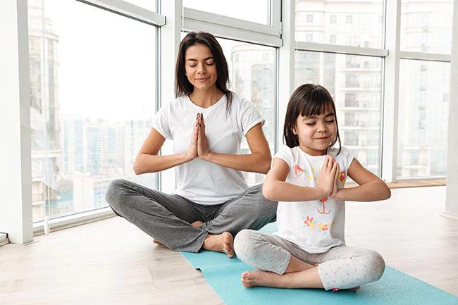Beautiful mother and child meditating at home sitting legs crossed on mat and doing yoga gesture with palms together