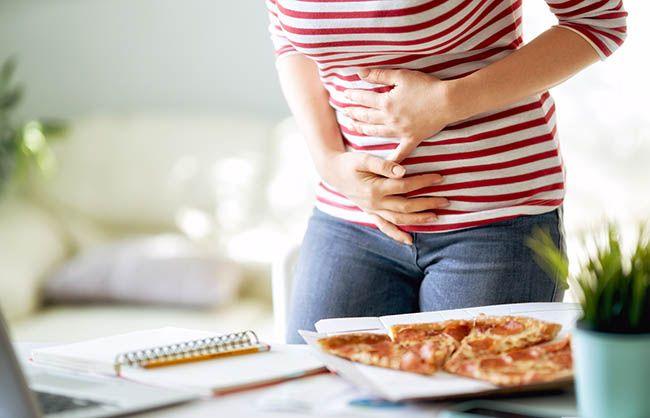 Woman eating processed pizza on the desk at work and having stomach problems
