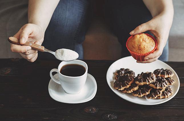 person adding sugar to coffee with one hand and holding sweet muffin in other