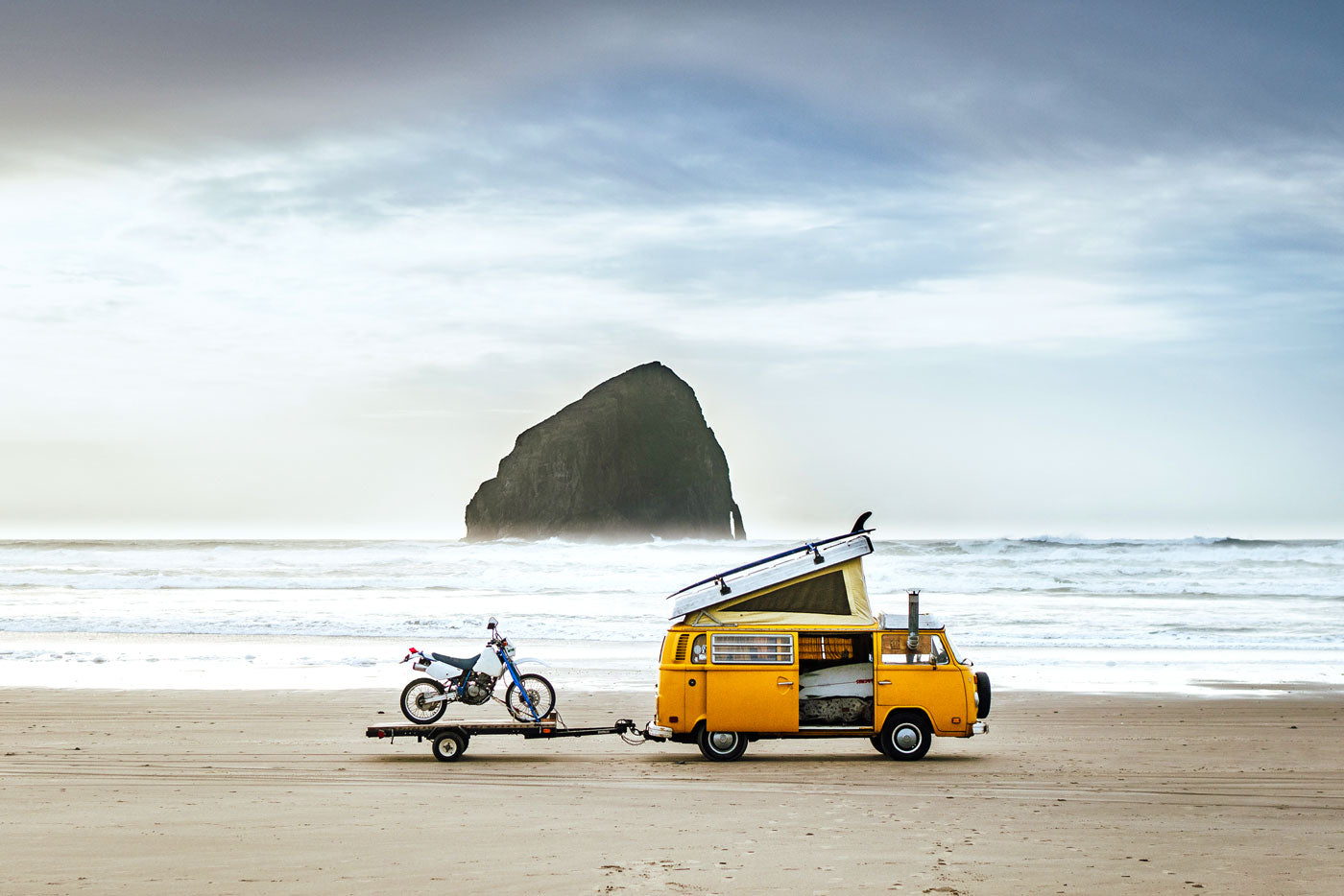 A van parked on a deserted beach. A motorcycle is parked on a hanger attached to the van. In the background is a rock formation in the ocean. (Photo: James Barkman)