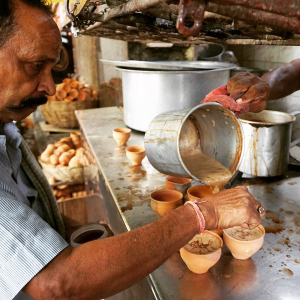 Cups of Chai tea being poured out in Kolkata, India. A very large pan that's being used to make chai is in the background, with a man helping to pour the tea in the foreground.