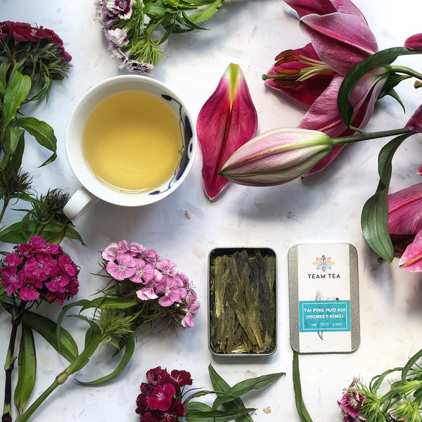 A cup of freshly brewed loose leaf tea, sat alongside an open tin of Team Tea's Tai Ping tea. The scene is beautiful set with a surrounding of fresh flowers.