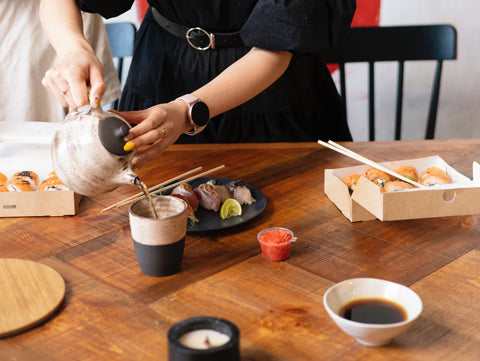 A lady pouring tea from a teapot into a cup surrounded by sushi and japanese food