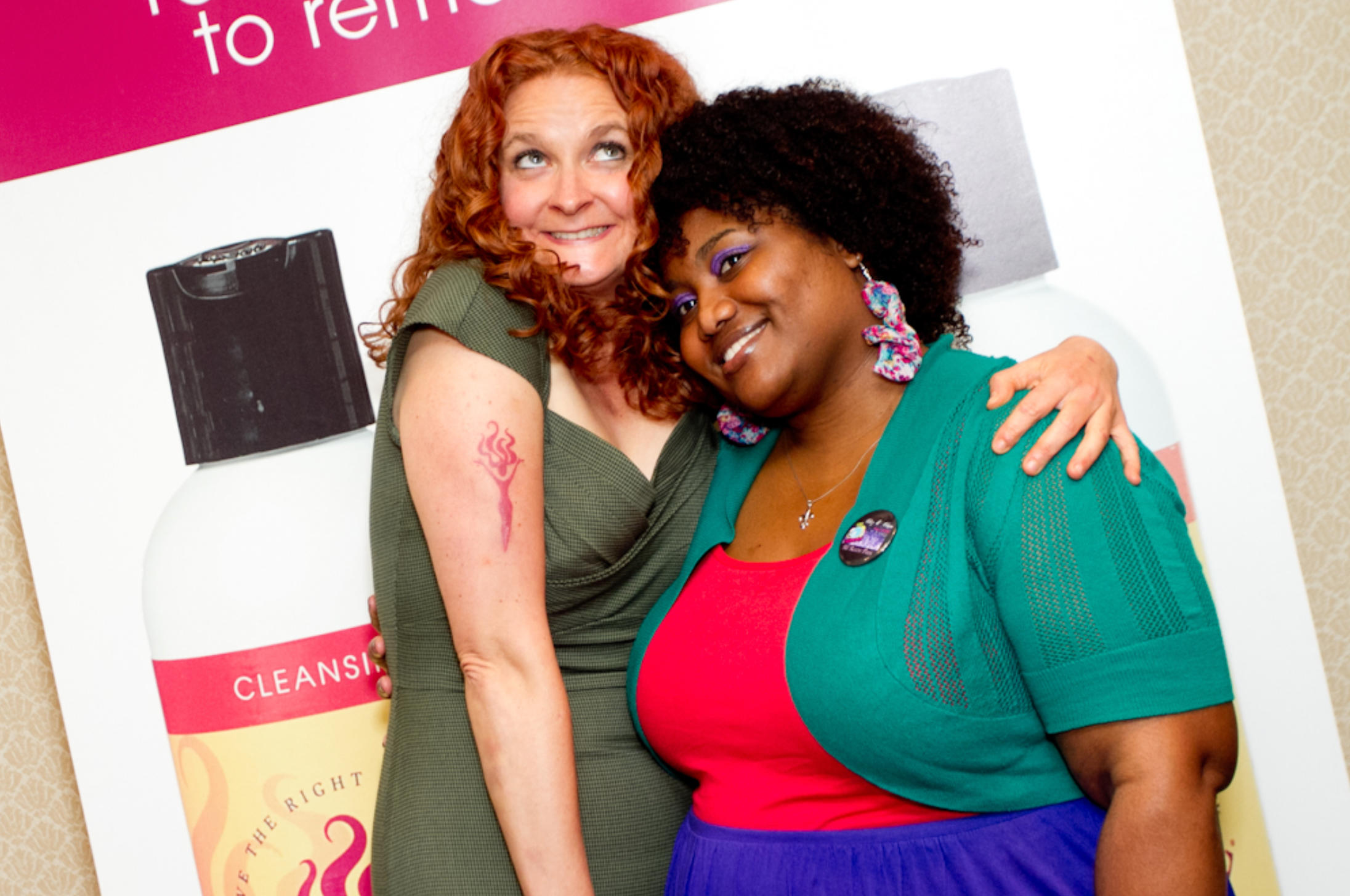 two curly haired women hugging and smiling for the camera in front of a poster