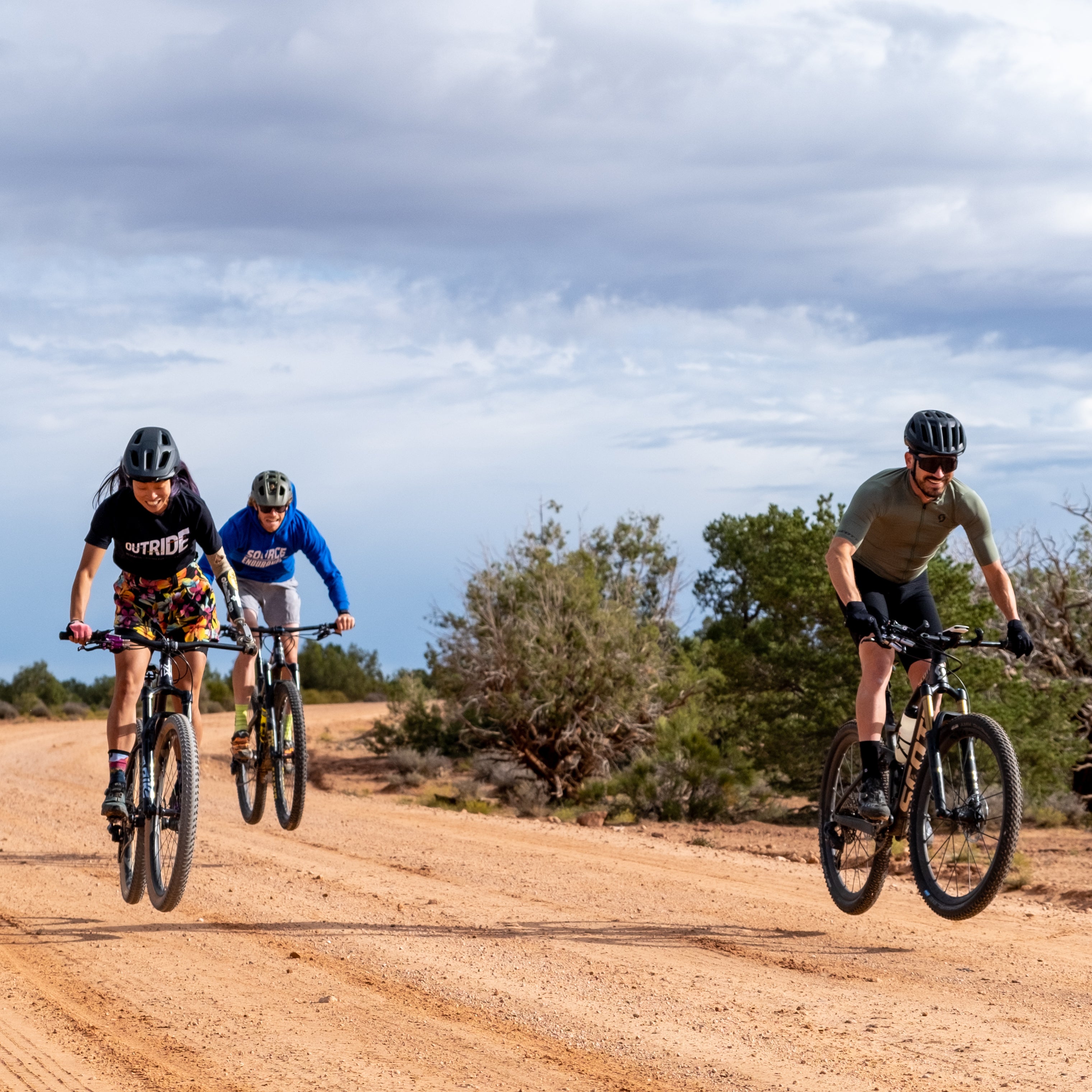Two adaptive mountain bikers ride side by side leading a third rider into a coordinated lift with the front and rear wheels level and in sync with each other. As a consequence, all three riders are also synchronizing their smiles.  Photo credit: Matt Didisheim https://www.instagram.com/didisheim/