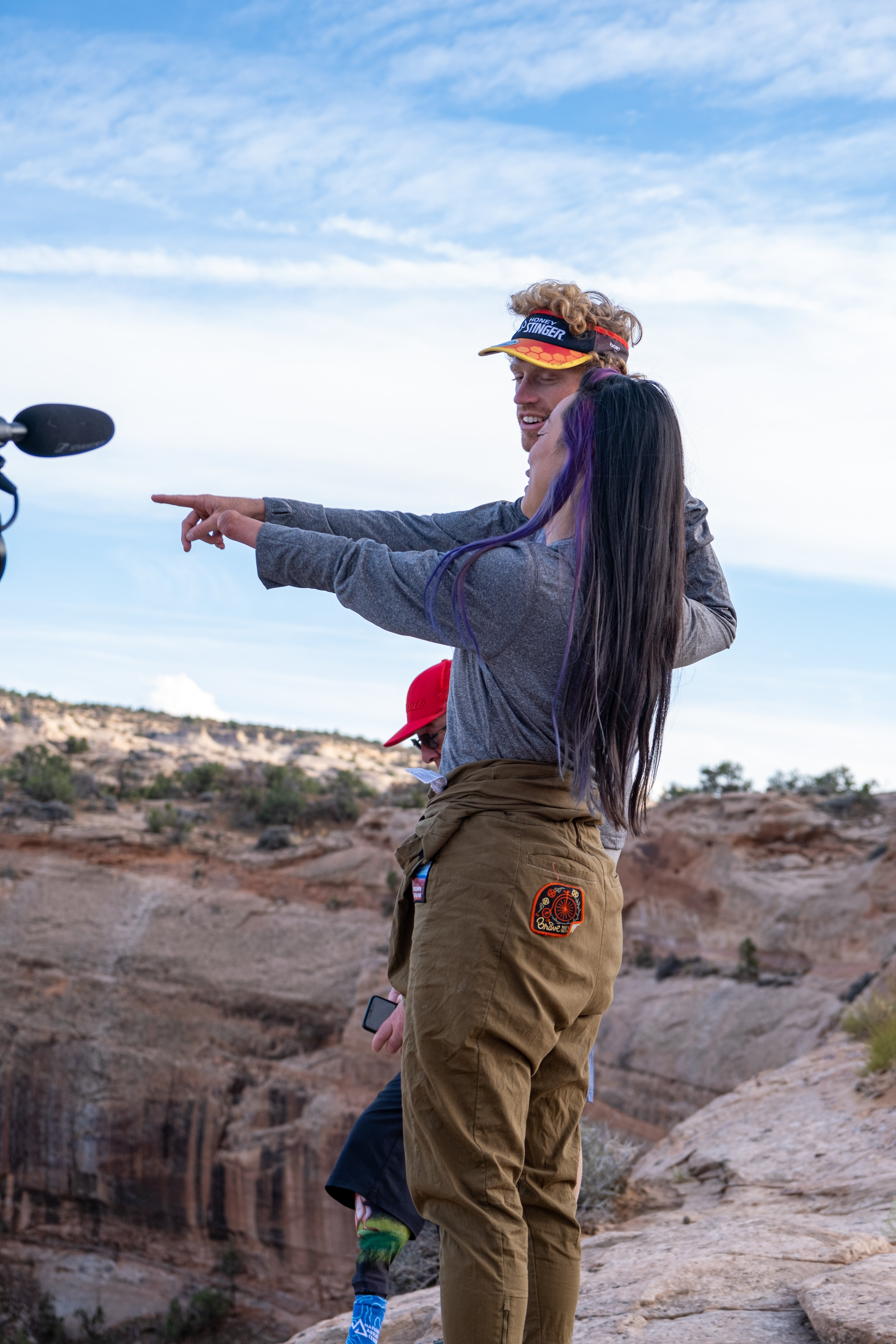 Josie and Taylor stand at the top edge of the Shafer climb in Canyonlands National Park with Roger in the background. Josie visualizes the finish line and points with her left residual limb while Taylor points with his right hand; their pointing converges making it seem like Taylor’s right hand could be Josie’s left hand upon first glance.  Photo credit: Matt Didisheim https://www.instagram.com/didisheim/