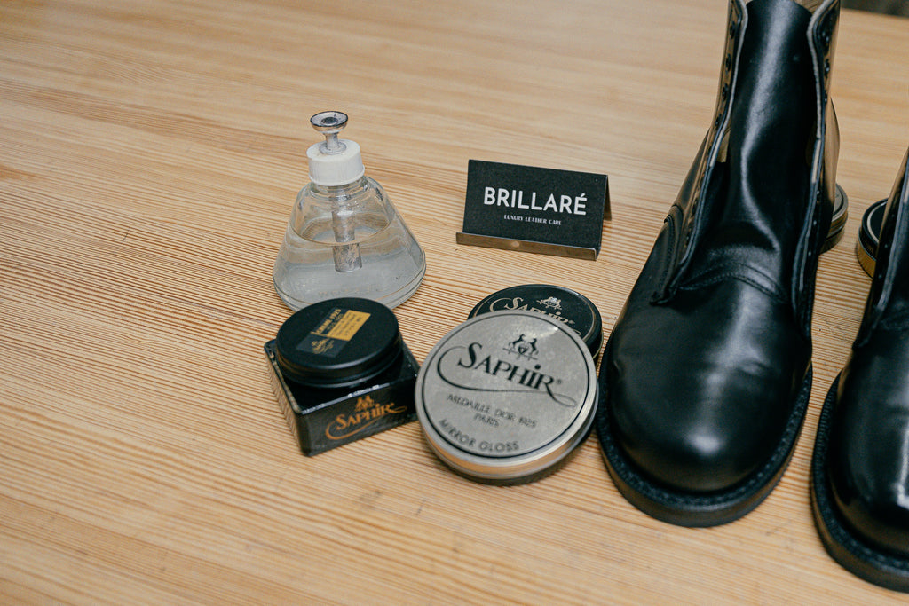 Up close image of prepping your station for a military parade boot mirror shine. Close up of brillaré business card, brillaré high shine water dispenser, Saphir Medaille d'or mirror gloss wax polish, saphir medaille d'or pate de luxe paste wax polish and military cadet boots, all in black