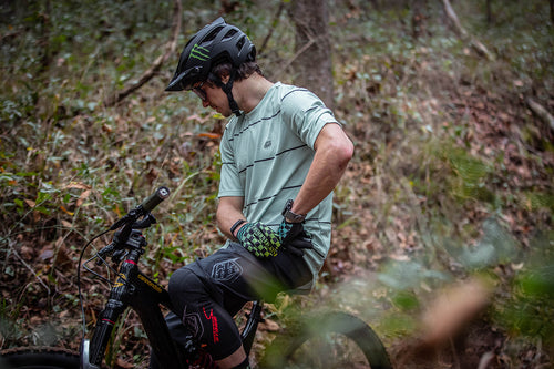 Male cyclist wresting on their bike wearing the Troy Lee Designs Flowline jersey, gloves, and shorts, and the TLD A3 MIPS helmet