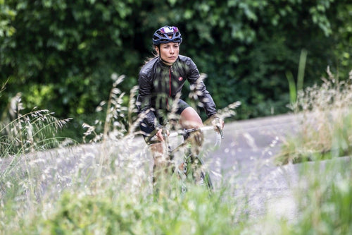 Female cyclist riding up a hill wearing the Castelli Idro 2 jacket. In the foreground is long grass in soft focus.