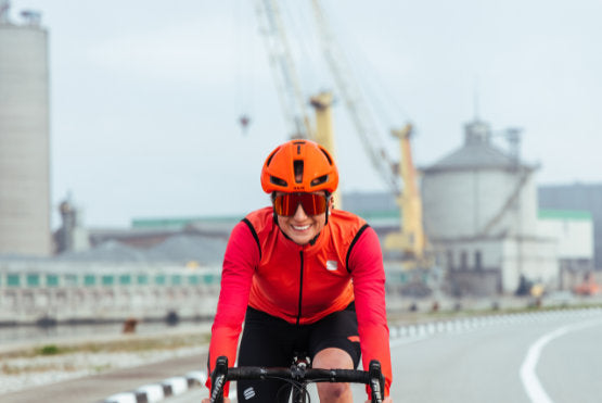 Smiling female cyclist riding towards the camera against an industrial backdrop wearing a raspberry Sportful BodyFit Pro Thermal Jersey and orange vest
