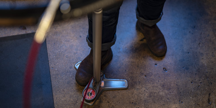 Silca Superpista Ultimate Floor Pump sitting on a workshop floor being anchored by a booted foot