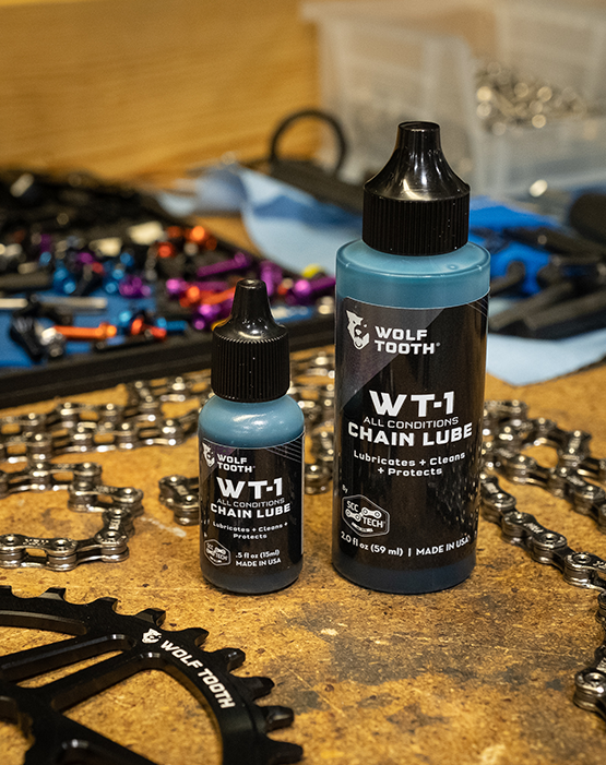 Two different sizes of bottle of blue Wolf Tooth WT-1 Chain Lube on a table full of tools and bike parts