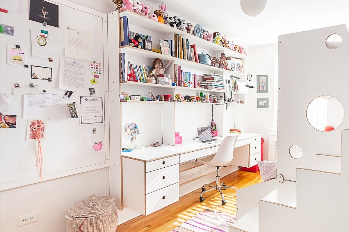 Bright study room with white furniture, shelves of books and toys, and a desk with papers.