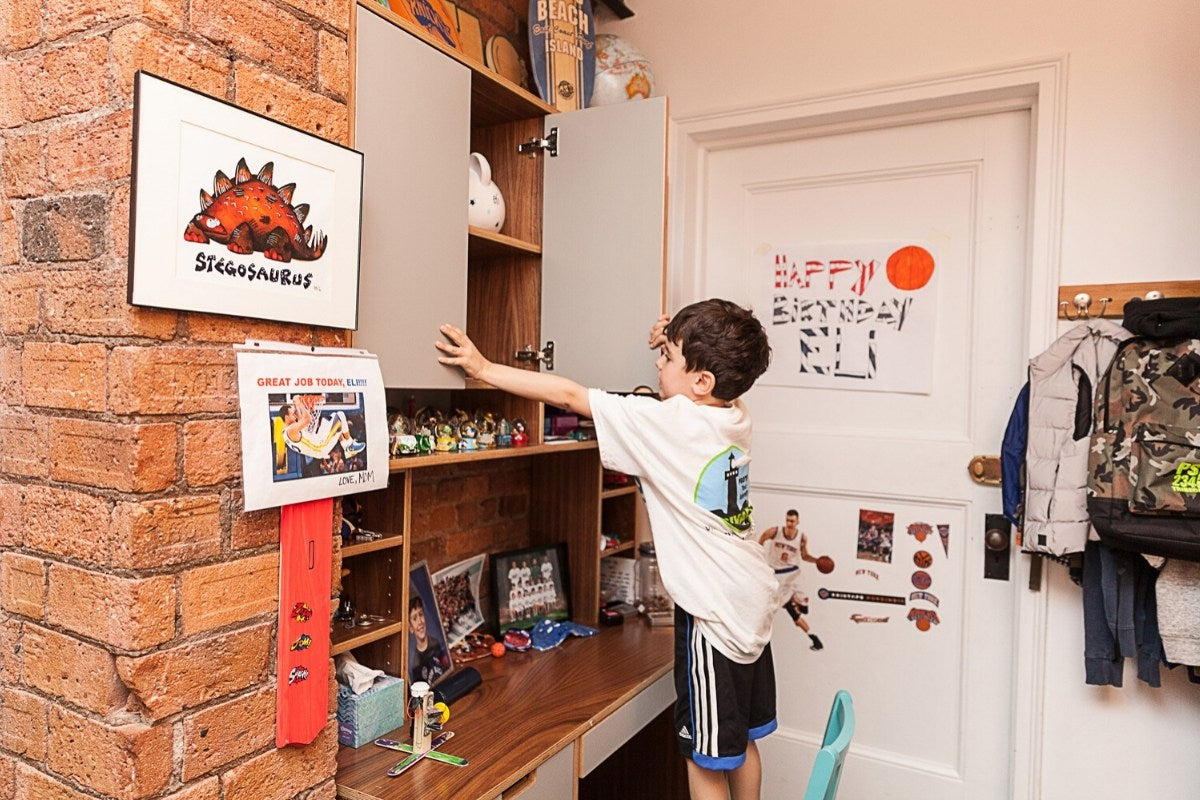 Child reaching for shelf with toys, books, and dinosaur picture.