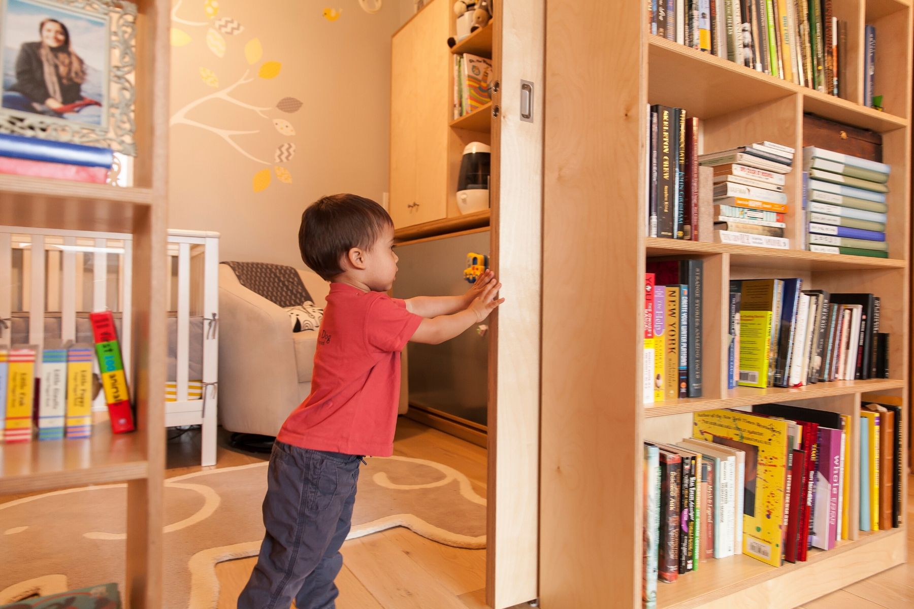 Toddler reaches for book on shelf, cozy room, yellow tree mural.