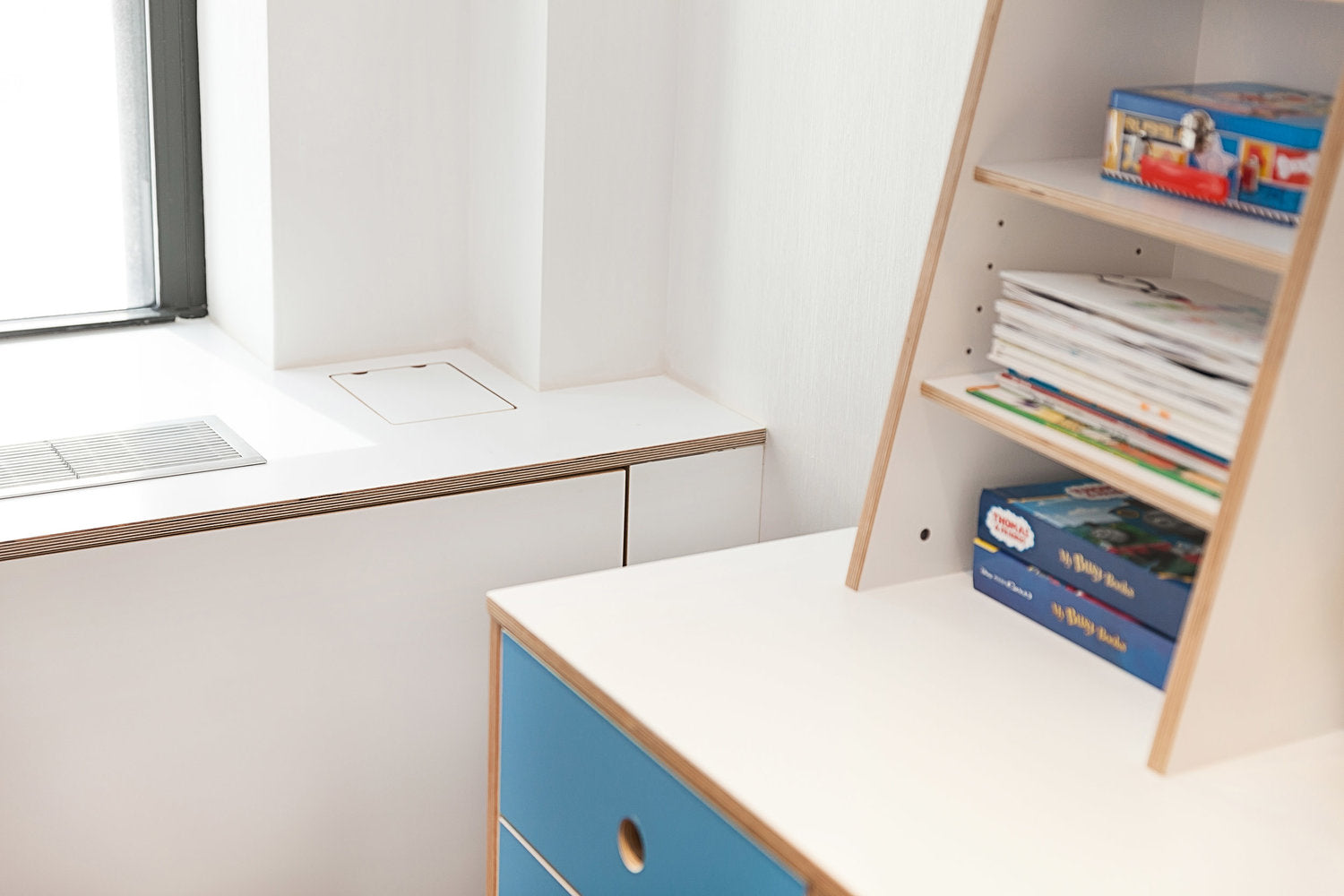 White desk, blue drawer, bookshelf with books and games in a bright room.