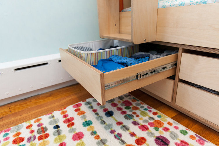 Open storage drawers under a bed showing organized compartments with clothes, in a room with floral rug.