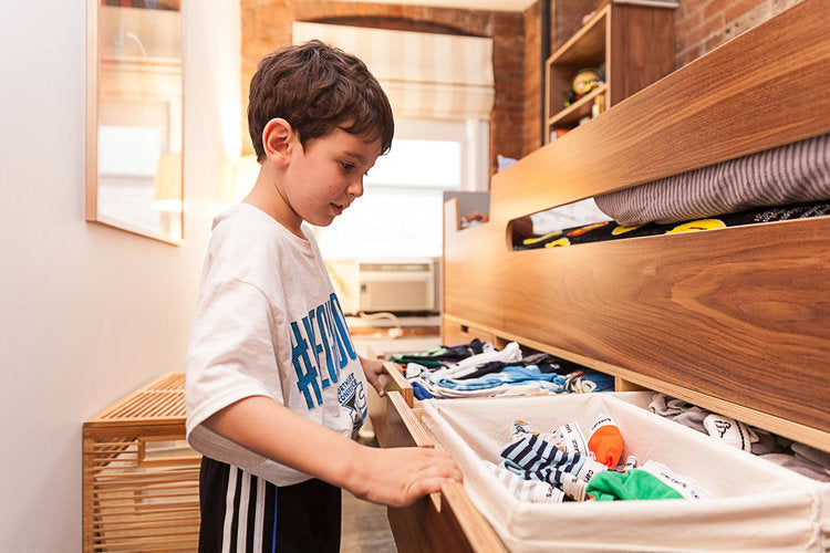 Young boy in a bedroom, pulling out a storage drawer under a bed, with clothes and toys inside.