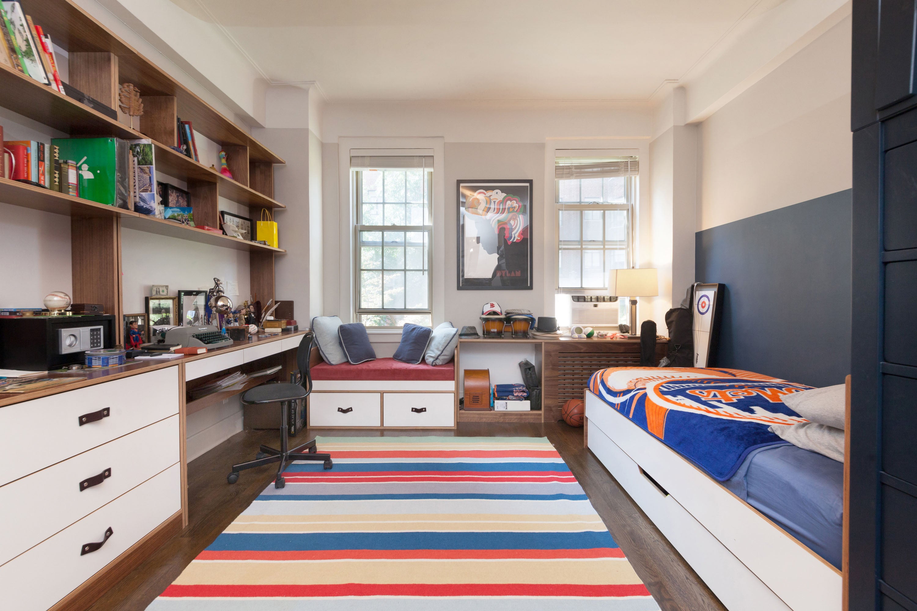 Modern bedroom with desk, bookshelves, striped rug, sports-themed bedspread.