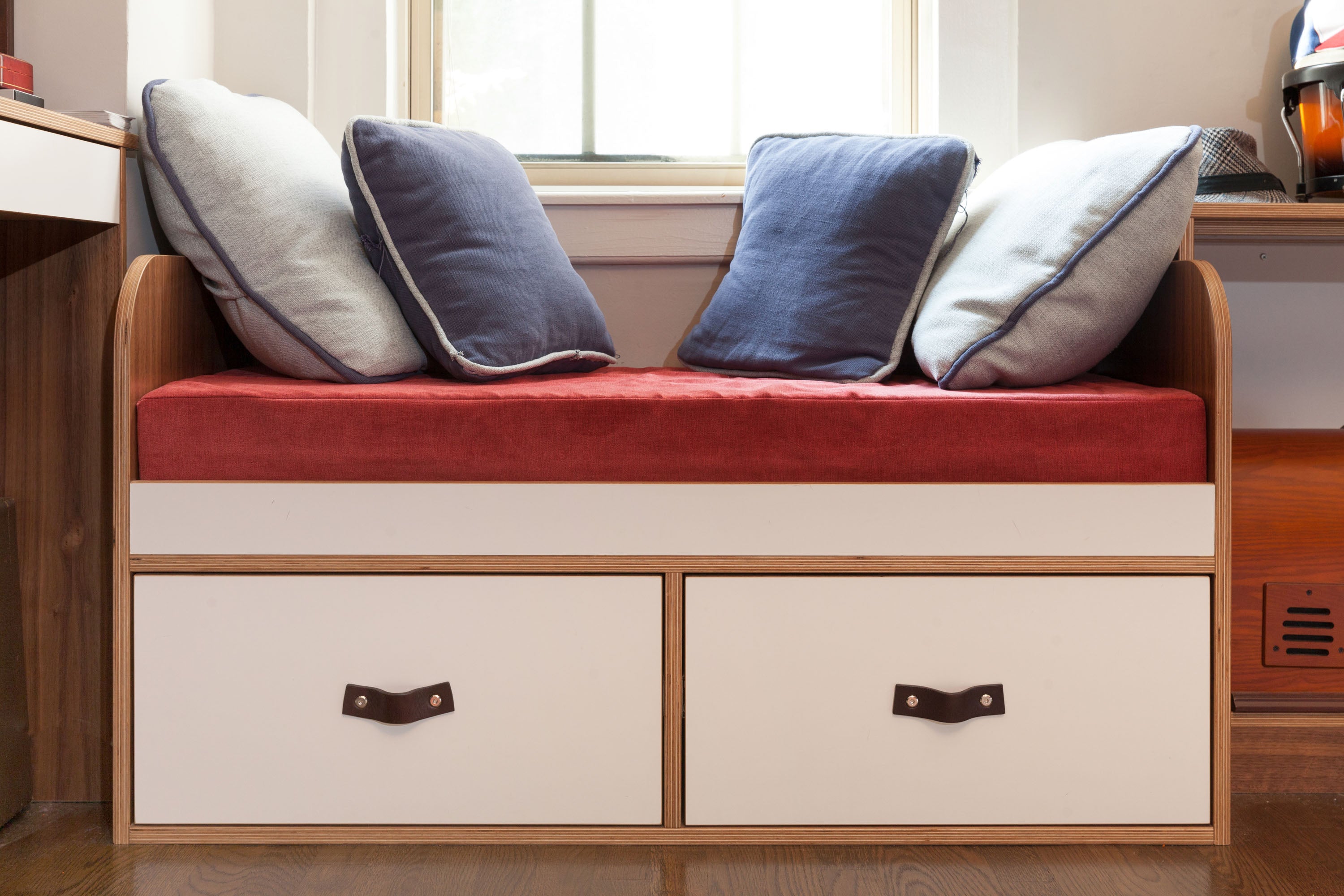 Window seat with red cushion, blue pillows, drawers, in a cozy nook.