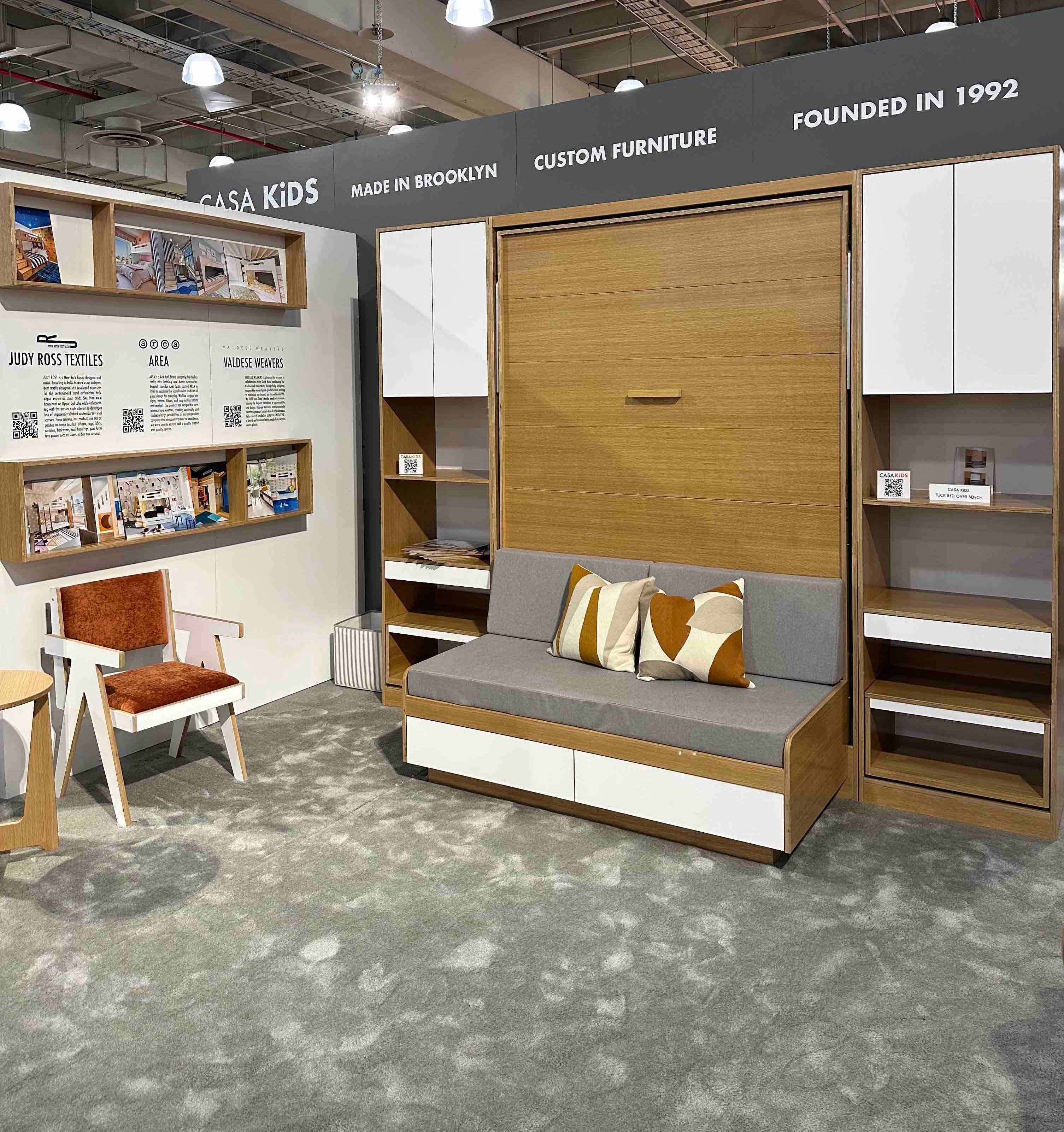 Casa Kids booth featuring a stylish murphy bed, sofa, chair, and wall-mounted shelving units at a fair.