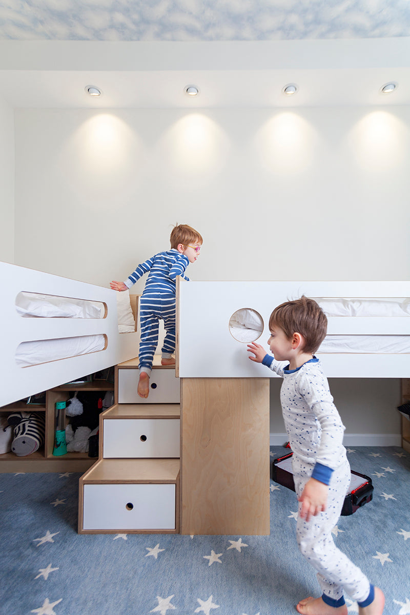 Children in pajamas play near bunk bed with stairs, drawers.