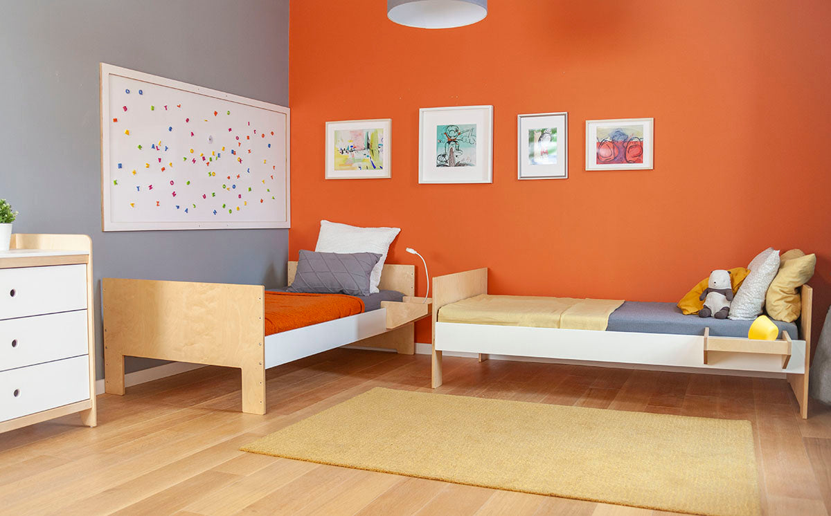 Bright children's room with twin beds, orange wall, artwork, and wooden furniture on a beige rug.
