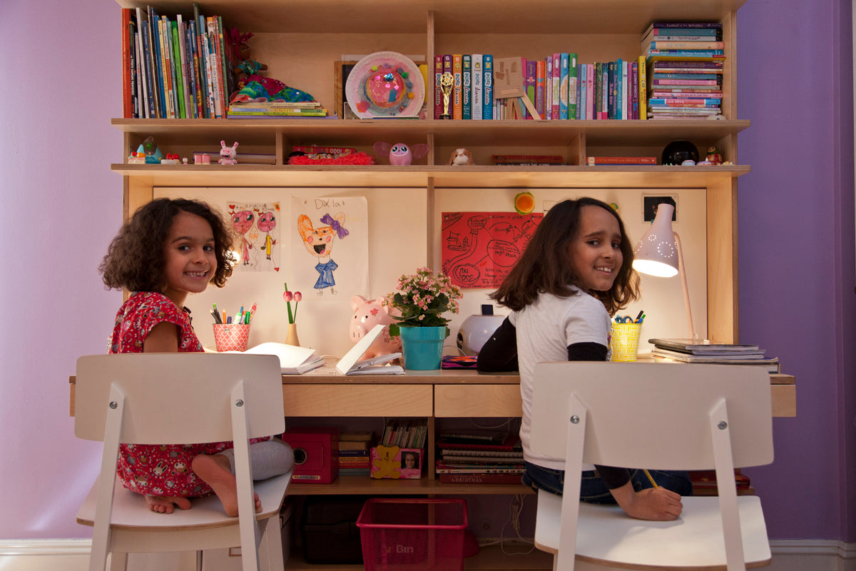 Two happy children studying at a desk with colorful bookshelves and creative drawings in the background.