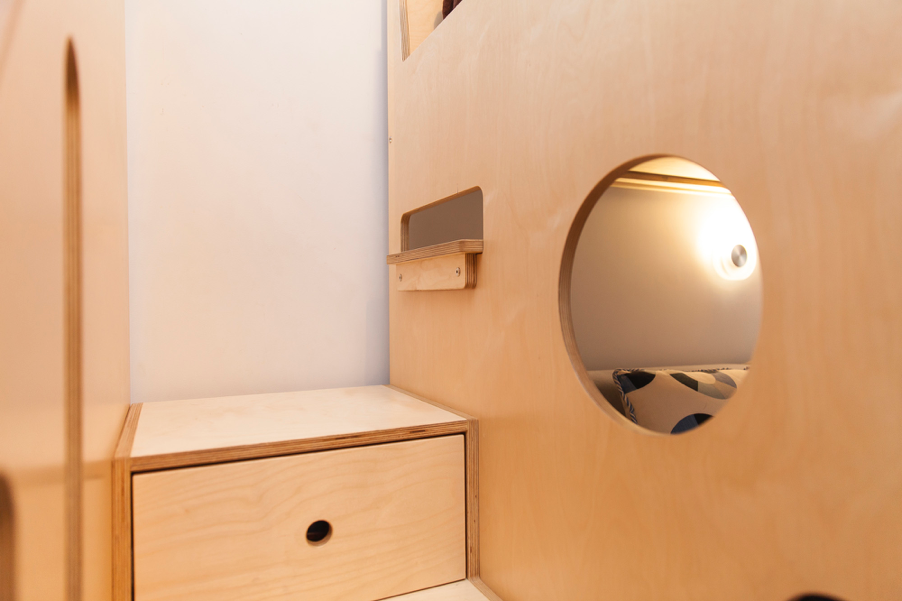 Circular window on wooden wall, bed with light reflection, bedside table.