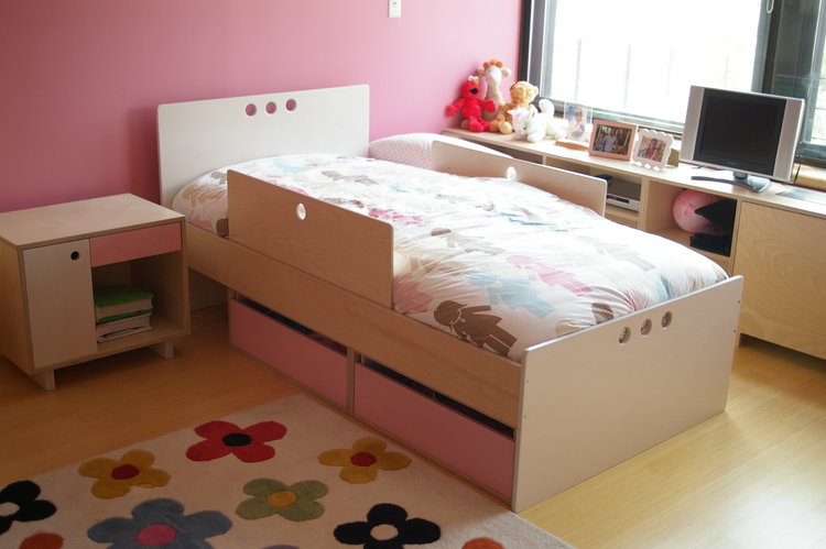 Bright pink children's room with a floral rug, bed with storage, nightstand, and a small desk.