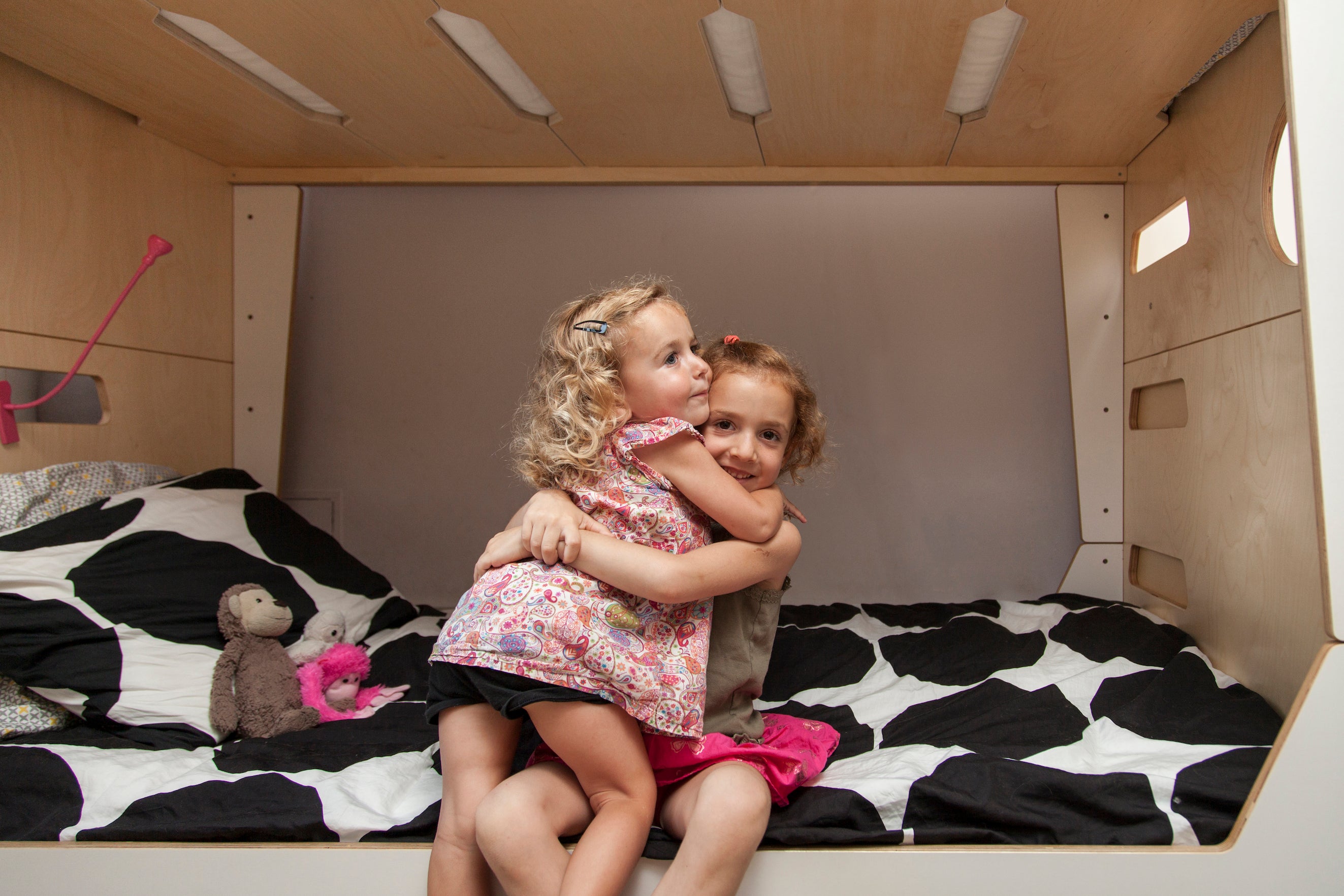 Two young girls hugging on a bed inside a cozy, wooden cabin-like space, with toys around them.