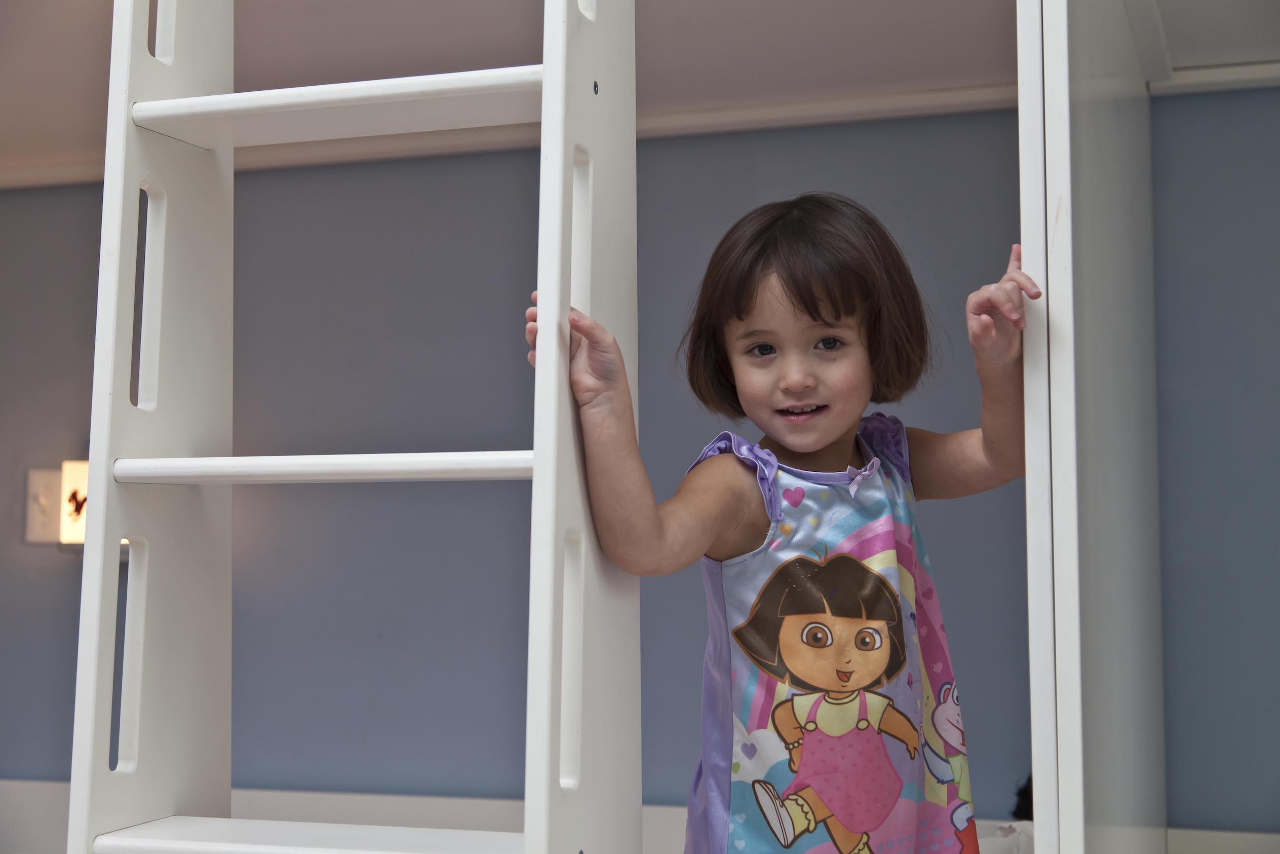 Young girl in a colorful dress smiling and holding onto white bunk bed ladders in a blue room.
