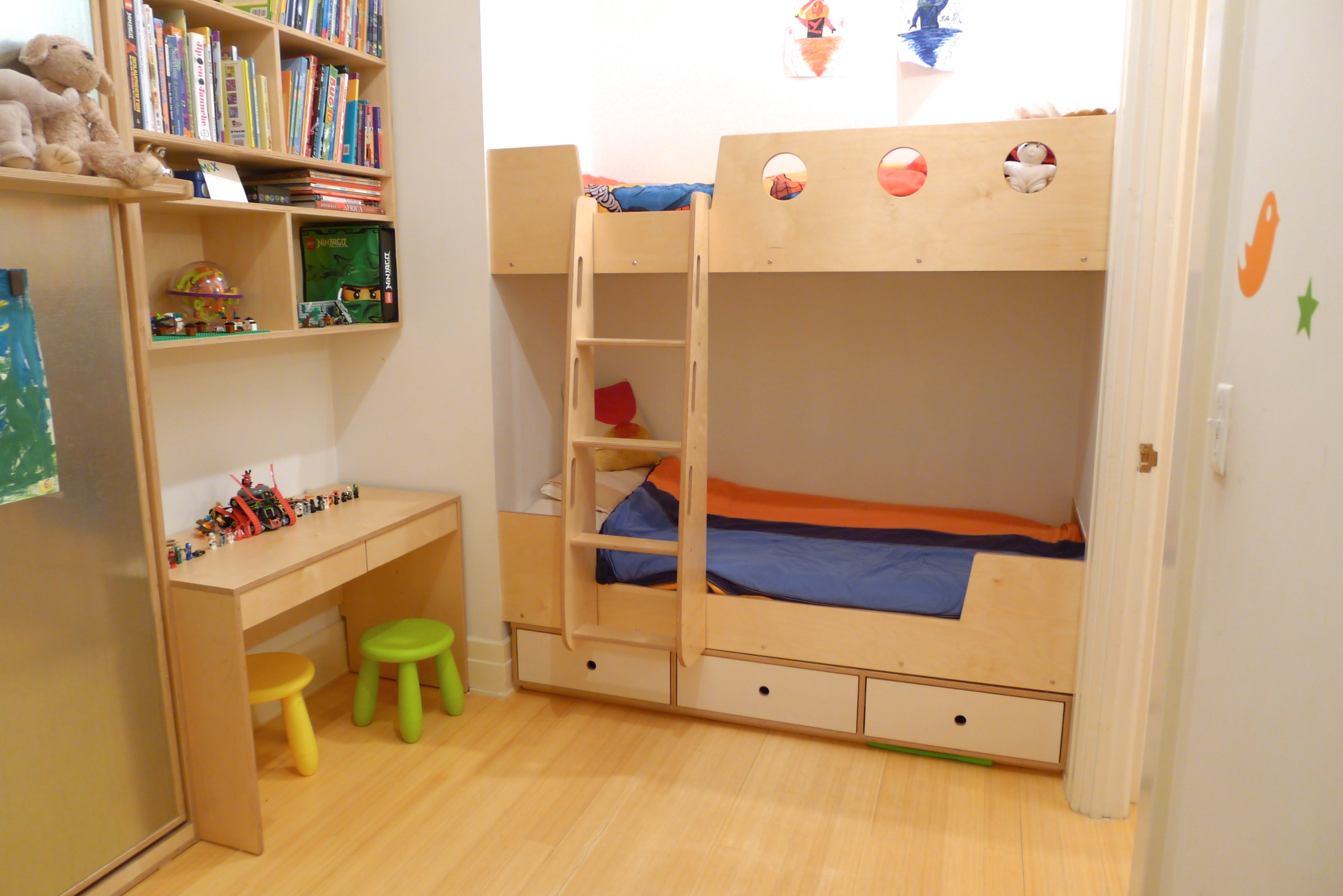 Child's bedroom with bunk beds, a small desk with stools, and a bookshelf filled with toys and books.