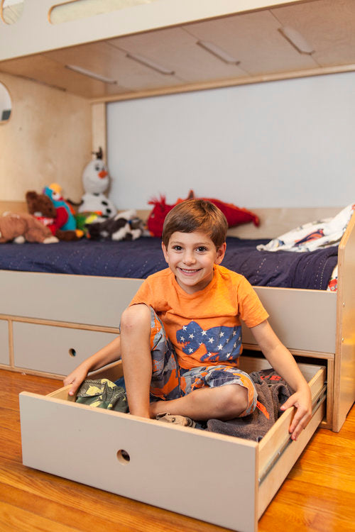 Young boy smiling and sitting in an open storage drawer under a bed surrounded by plush toys.