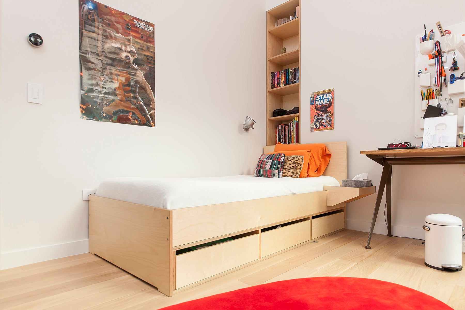 Minimalist child's bedroom with single bed, desk, artwork on wall, and red circular rug.