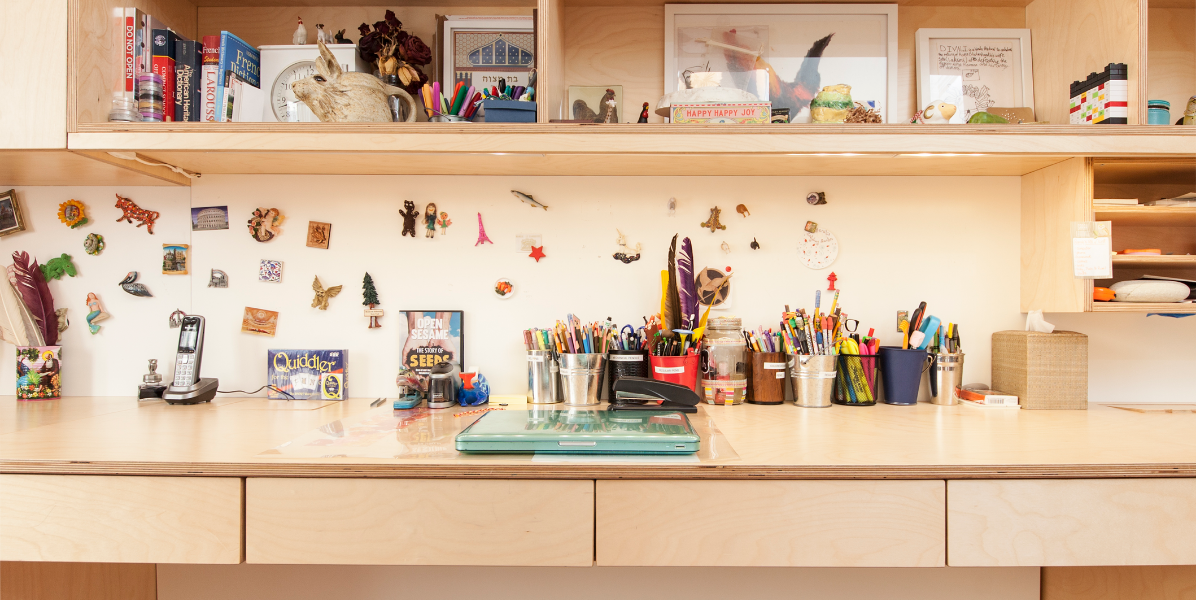 A tidy desk with art supplies, books, and decorations, including various magnets on the wall and items on the shelves.
