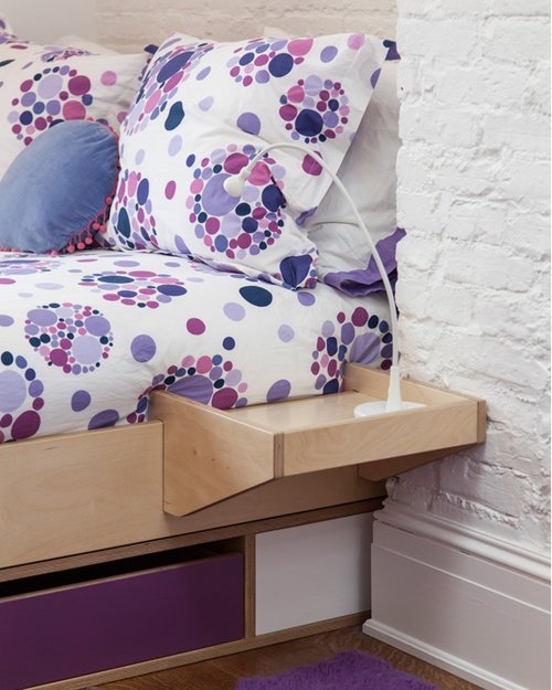 Detail of a bed with colorful polka-dot bedding and a wooden bedside shelf against a brick wall.